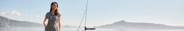 Elegante Frau am See mit Segelboot, stilvolle Mode, entspannte Sommeratmosphäre.