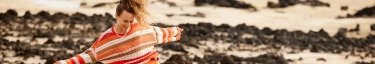 Lebensfreude am Strand: Frau in buntem Pullover tanzt auf Felsen.